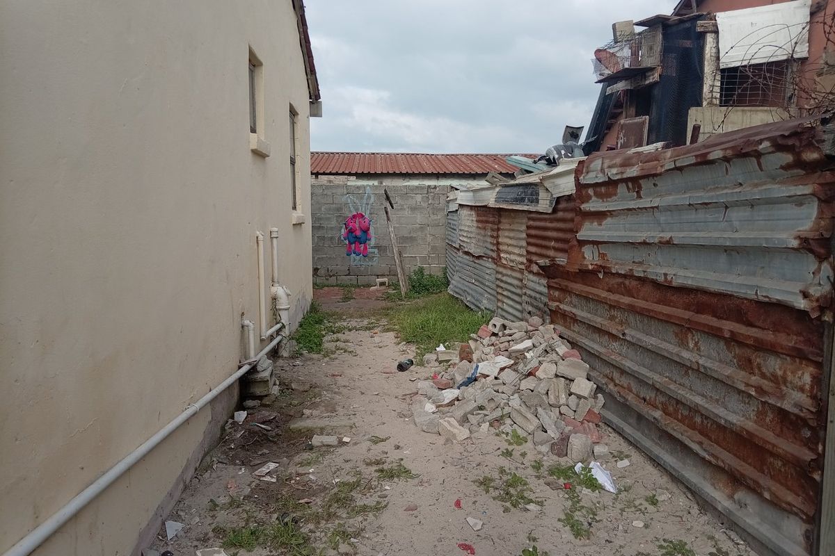 A narrow side or rear exterior passage featuring exposed plumbing, a concrete block boundary wall, and weathered corrugated metal fencing with significant debris.