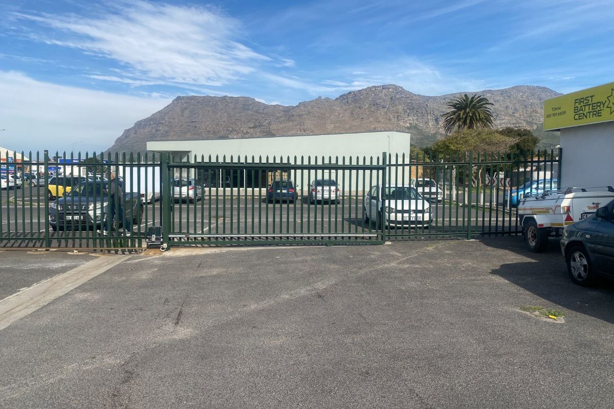 Front view of the industrial property featuring a secure steel palisade fence, sliding gate, and paved parking area with mountain views.