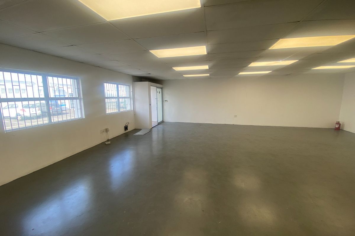An open-plan industrial office space featuring concrete flooring, suspended ceiling with recessed lighting, and security bars on the windows.
