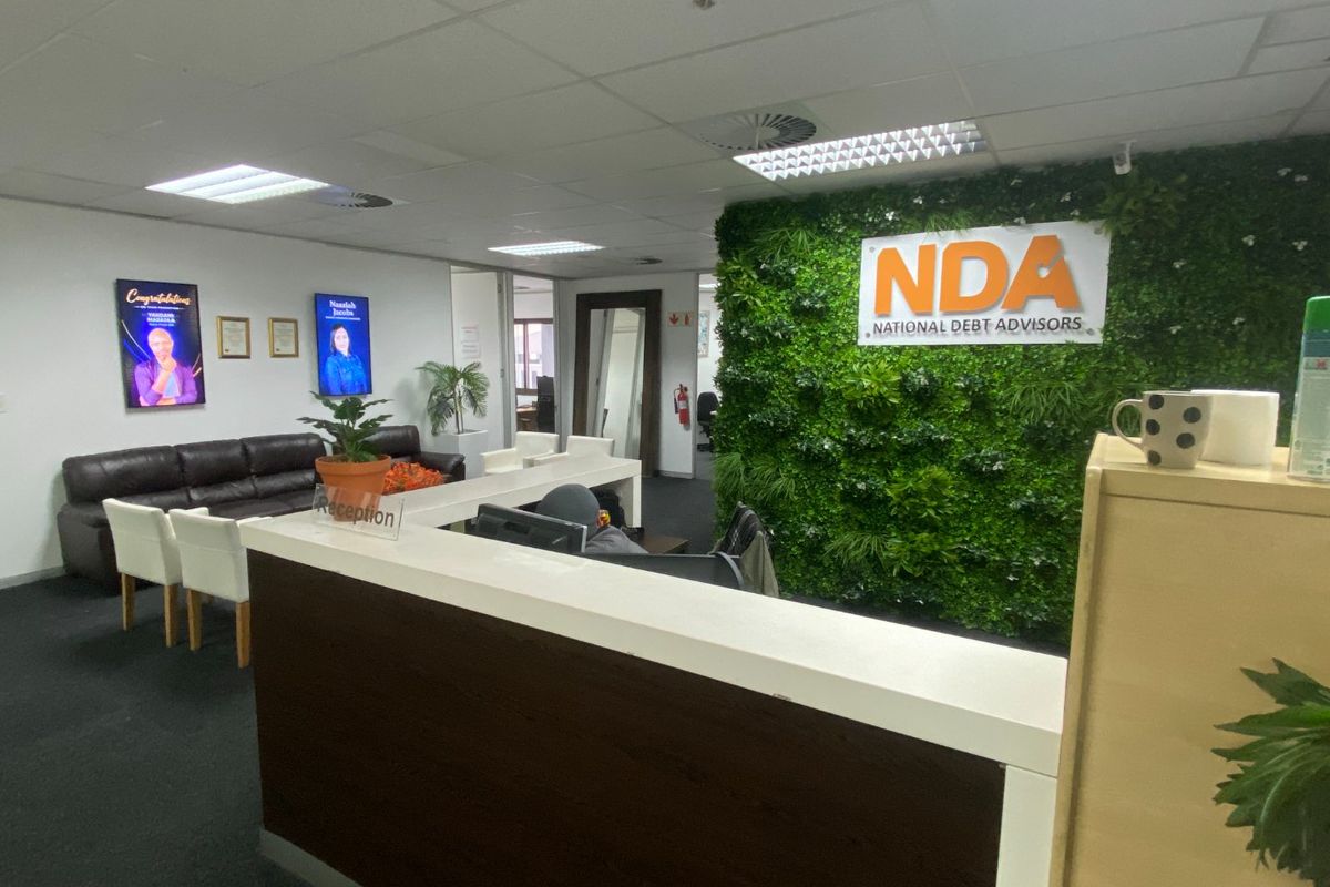 Commercial office reception area featuring a built-in reception desk, suspended ceiling with recessed lighting, and a prominent branded artificial green wall.