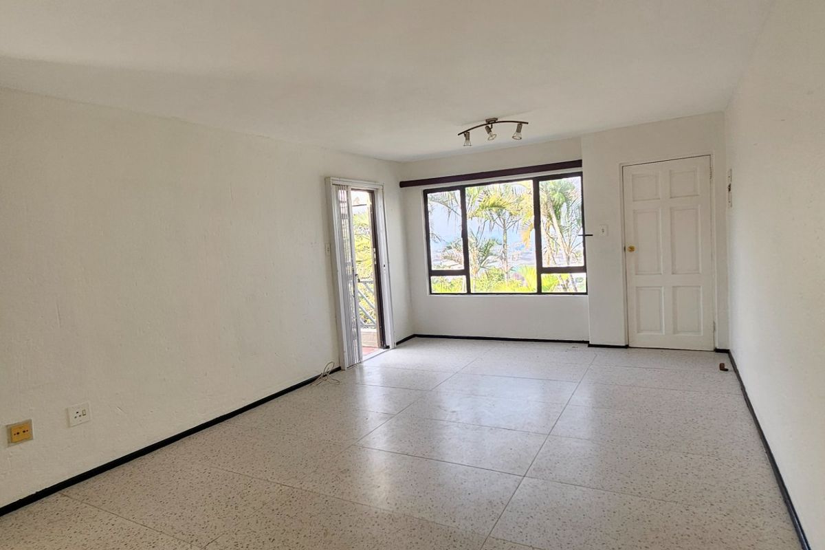 An empty, neutrally painted living room featuring tiled flooring, a large window with natural light, and access to an outdoor area via a sliding door.