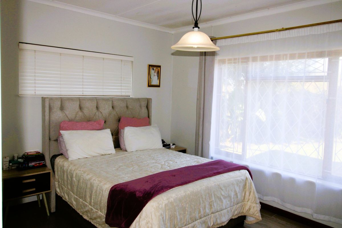 A well-lit bedroom featuring a large window with security bars, venetian blinds, and a central pendant light fixture.