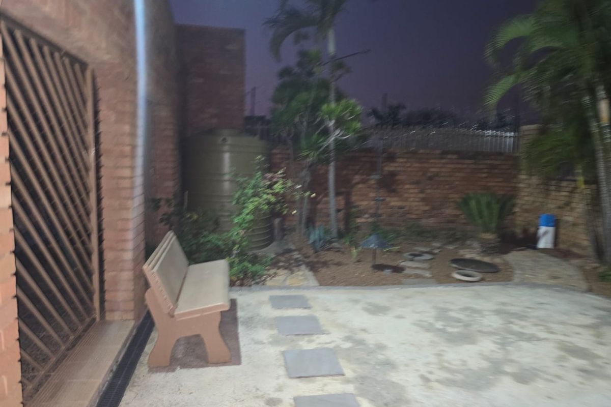 A rear exterior view of a brick house featuring a paved patio area, security gate, and a water tank in the garden.