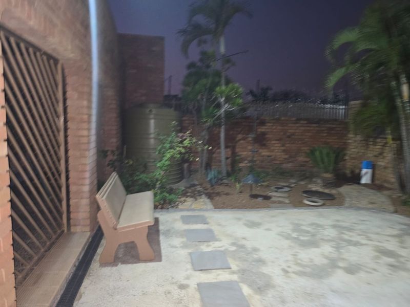 A rear exterior view of a brick house featuring a paved patio area, security gate, and a water tank in the garden.