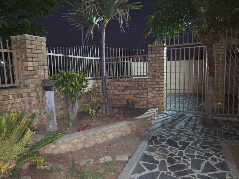 A night view of the property entrance featuring a brick boundary wall, metal security gate, and stone paved walkway with landscaped garden beds.