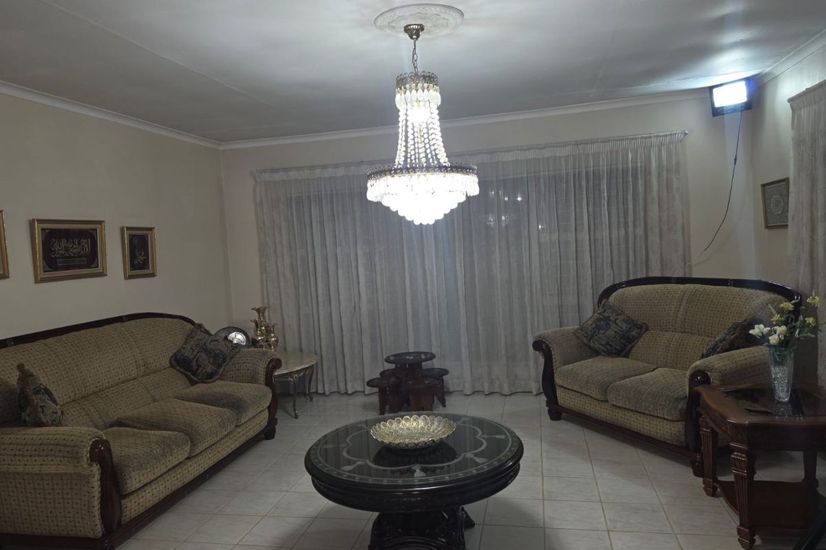 A traditional living room featuring tiled flooring, a central crystal chandelier with a decorative ceiling rose, and floor-to-ceiling window treatments.