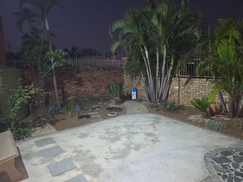 A private rear garden area featuring a concrete patio, brick boundary walls with security fencing, and established palm trees.