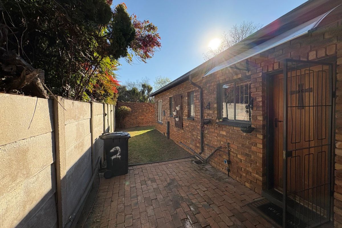 A rear exterior view of the townhouse featuring a brick-paved patio area, a small lawn, and a secure entrance with a metal gate.