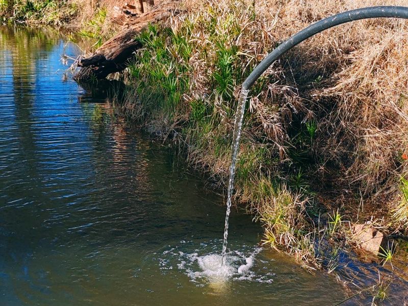 A natural farm pond featuring a water supply pipe feeding into the water body, set within a rural landscape.
