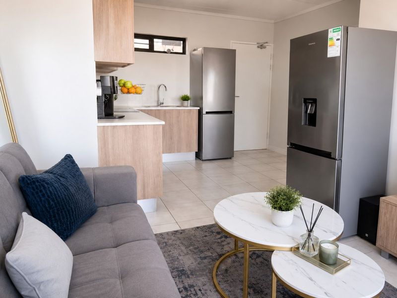 Modern open-plan living area featuring a compact kitchen with light wood-finish cabinetry, stone-look countertops, and neutral tiled flooring throughout.