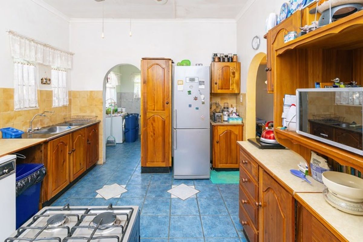 A dated kitchen featuring wooden cabinetry, tiled flooring, and a gas hob, with an adjacent scullery area accessible through an archway.