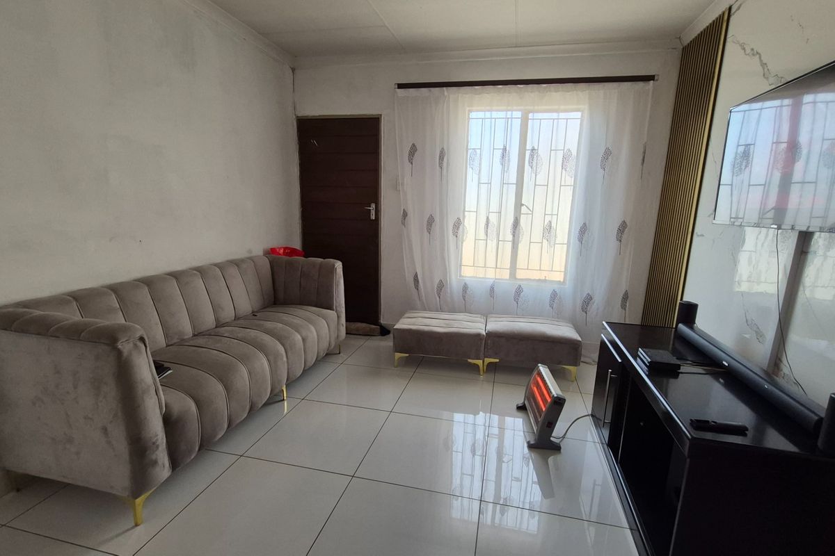 A living room featuring tiled flooring, decorative wall paneling, and a window with security bars. The walls show signs of uneven finish and staining.