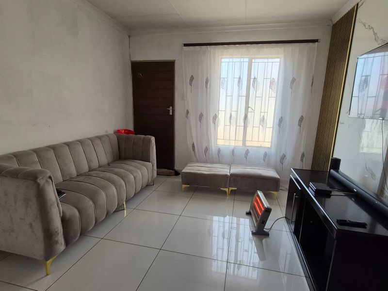 A living room featuring tiled flooring, decorative wall paneling, and a window with security bars. The walls show signs of uneven finish and staining.
