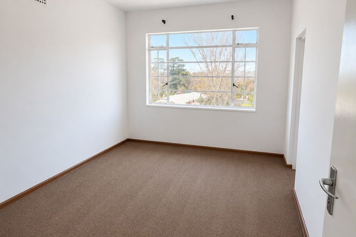 A bright, empty bedroom featuring neutral carpeted flooring, a large window with natural light, and a simple ceiling light fixture.