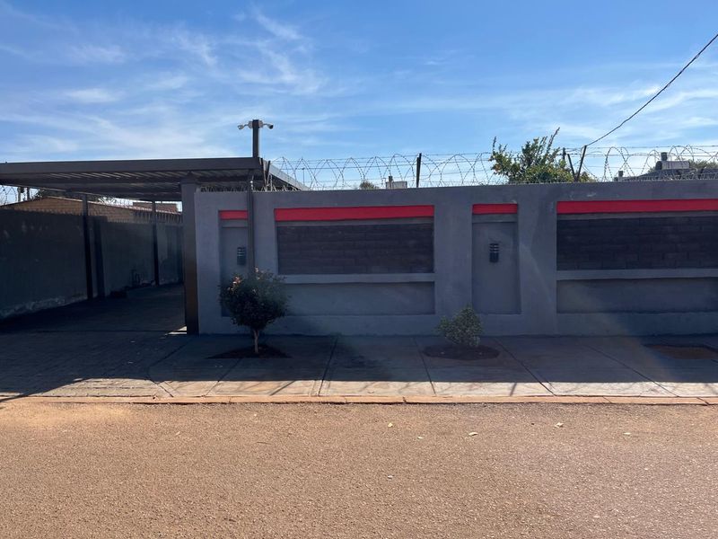 Front view of the property featuring a secure perimeter wall with razor wire, a paved driveway, and a covered carport area.