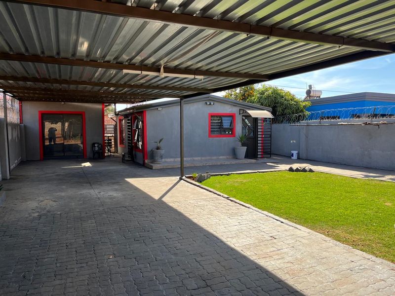 Rear exterior view featuring a large covered carport, paved driveway, manicured lawn, and secure perimeter walling with razor wire.