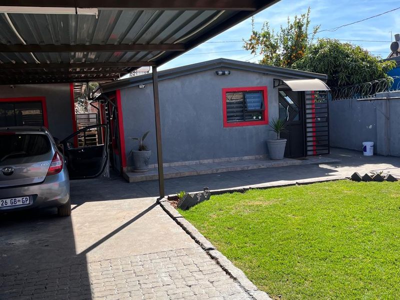 Rear exterior view featuring a paved driveway, a covered carport, a small lawn, and a secure boundary wall with razor wire.