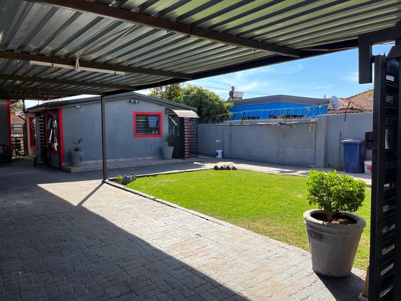 Rear exterior view featuring a paved driveway, covered carport, small lawn area, and perimeter wall with security fencing.