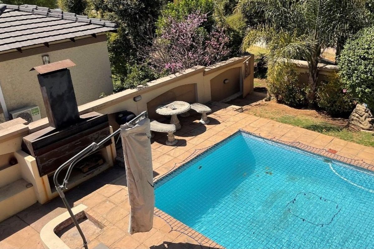 Outdoor pool area featuring a tiled patio, built-in braai, and a safety net-covered swimming pool, set against a landscaped garden boundary.