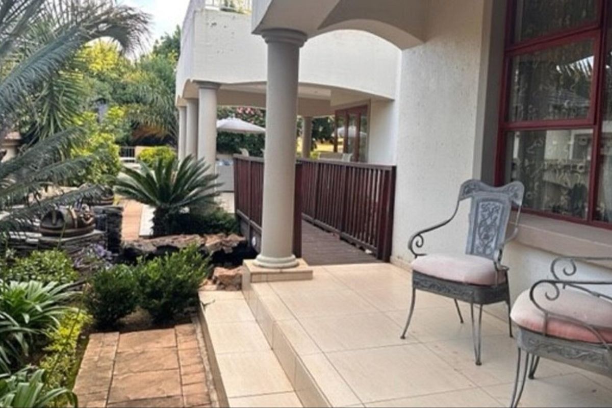 A covered patio area featuring tiled flooring and structural columns, overlooking a landscaped garden with a small wooden bridge.