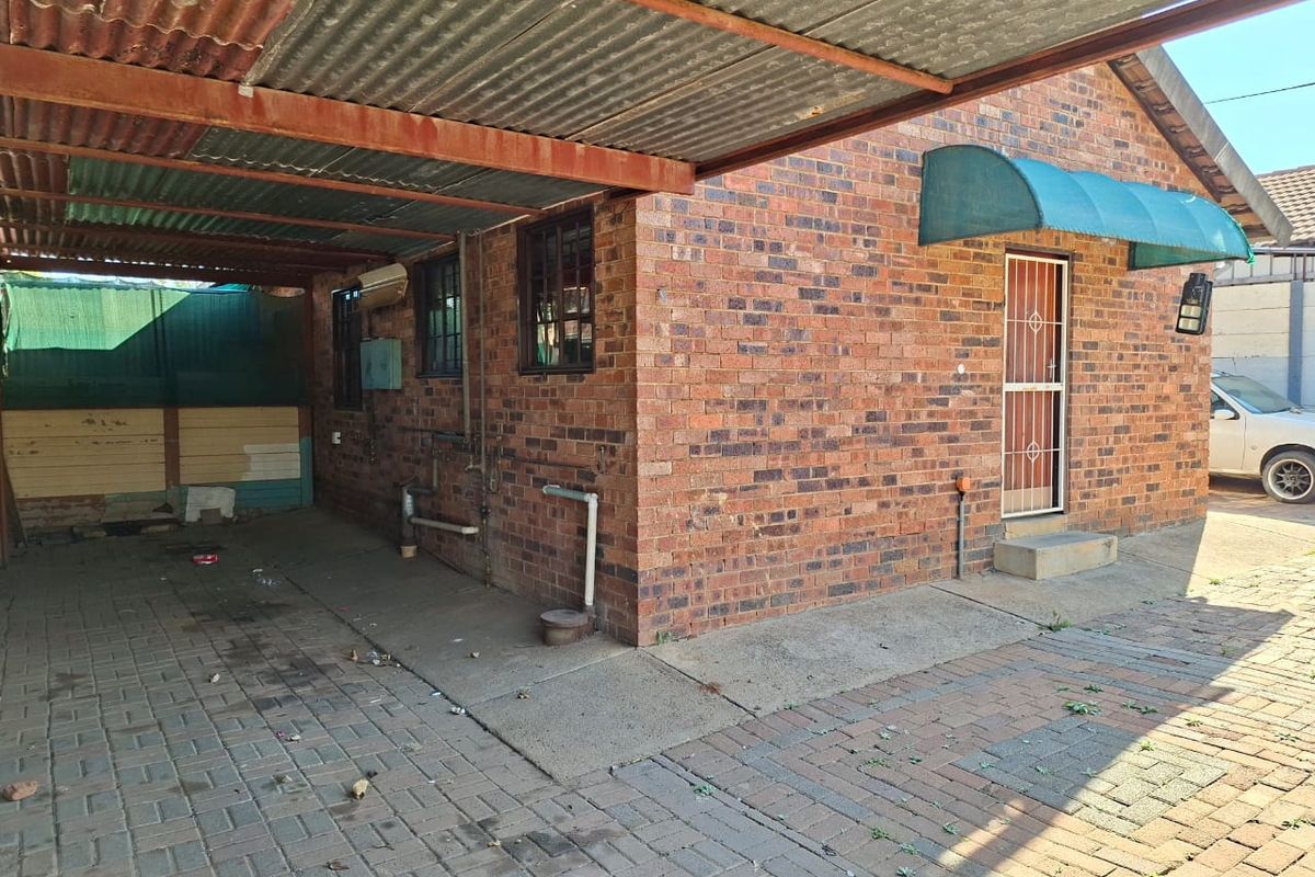 Rear exterior view featuring a brick facade, a corrugated iron carport, and paved driveway area with a security gate entrance.