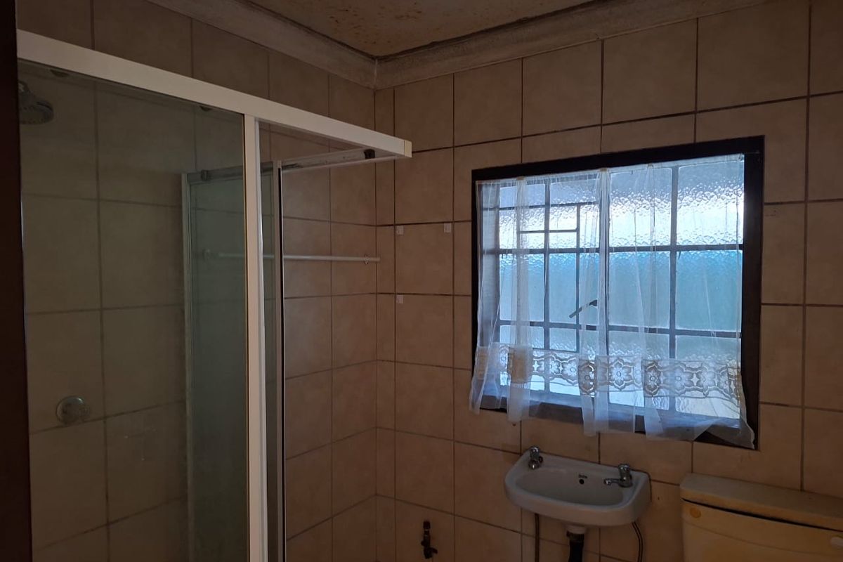 A dated bathroom featuring a shower enclosure, wall-mounted basin, and toilet, with tiled walls and a window with security bars.