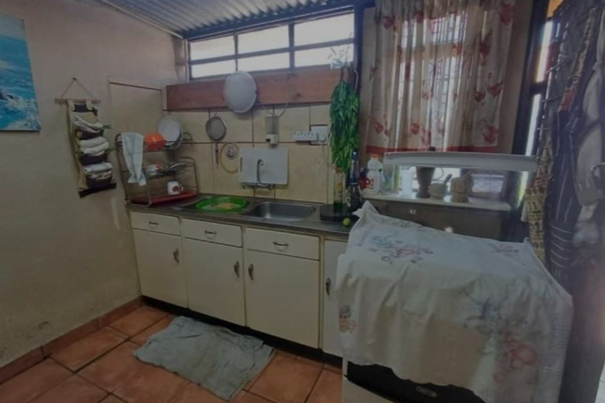 A functional kitchen area featuring built-in cabinetry, a stainless steel sink, tiled splashback, and terracotta flooring under a corrugated metal ceiling.