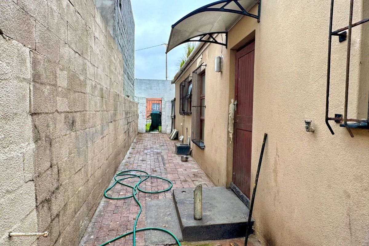 A narrow rear exterior walkway featuring brick paving, a concrete boundary wall, and a side entrance with a protective awning.