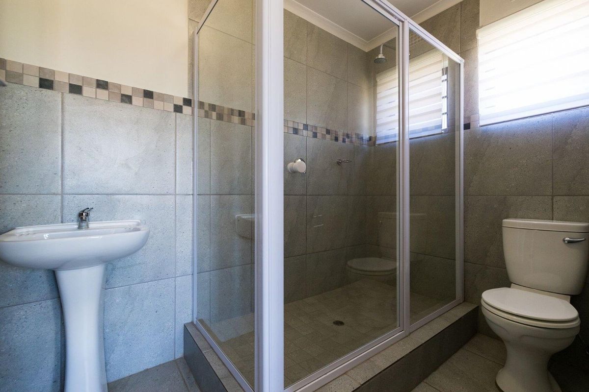 A clean, functional bathroom featuring a pedestal basin, a glass-enclosed shower, and a toilet, finished with neutral grey wall and floor tiles.