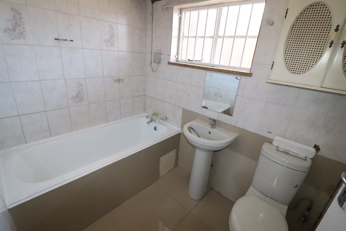 A dated bathroom featuring a standard bathtub, pedestal basin, and toilet, with tiled finishes and a window fitted with security bars.