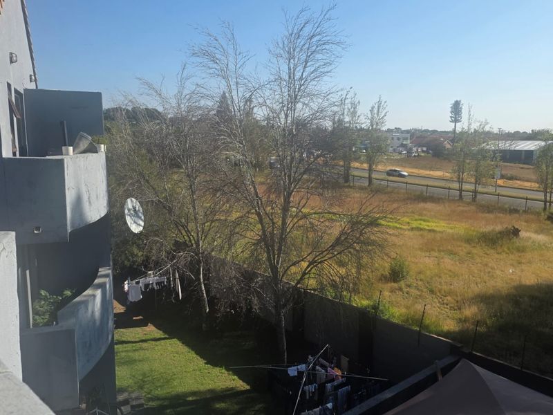 Elevated view from an apartment balcony overlooking a garden area, perimeter wall, and adjacent open land with a road in the distance.