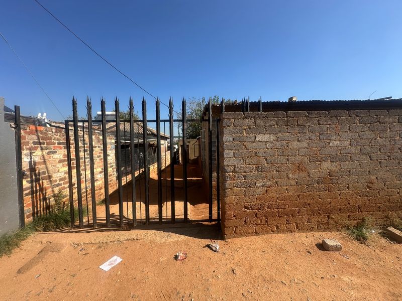 Front exterior view showing a metal spike fence, gated entrance, and brick boundary wall on an unpaved lot.