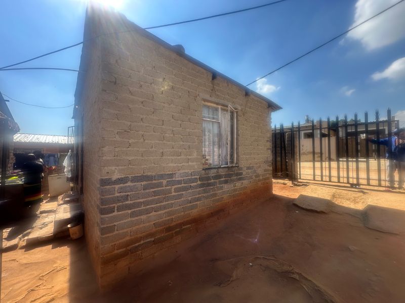 Exterior view of a modest residential structure featuring exposed brick walls, security window bars, and a metal perimeter gate.