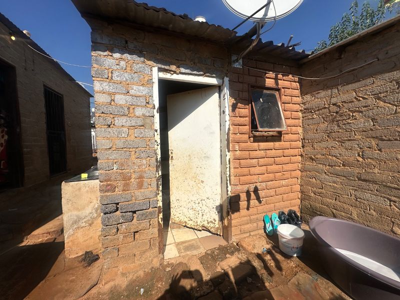 Exterior view of a small brick structure with a corrugated metal roof, showing significant wear to the masonry and door frame.