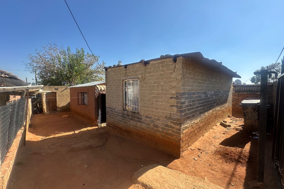 Exterior view of a modest residential structure featuring exposed brick walls and a corrugated metal roof set on an unpaved plot.