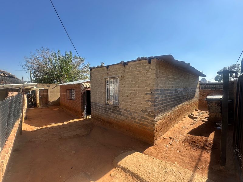 Exterior view of a modest residential structure featuring exposed brick walls and a corrugated metal roof set on an unpaved plot.
