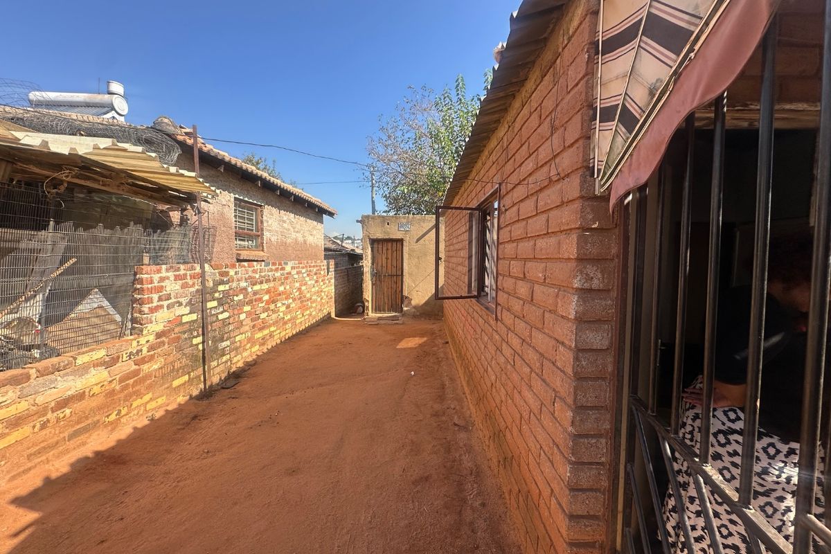 A side passage or rear exterior area featuring exposed brick walls, security bars on windows, and an unpaved ground surface.