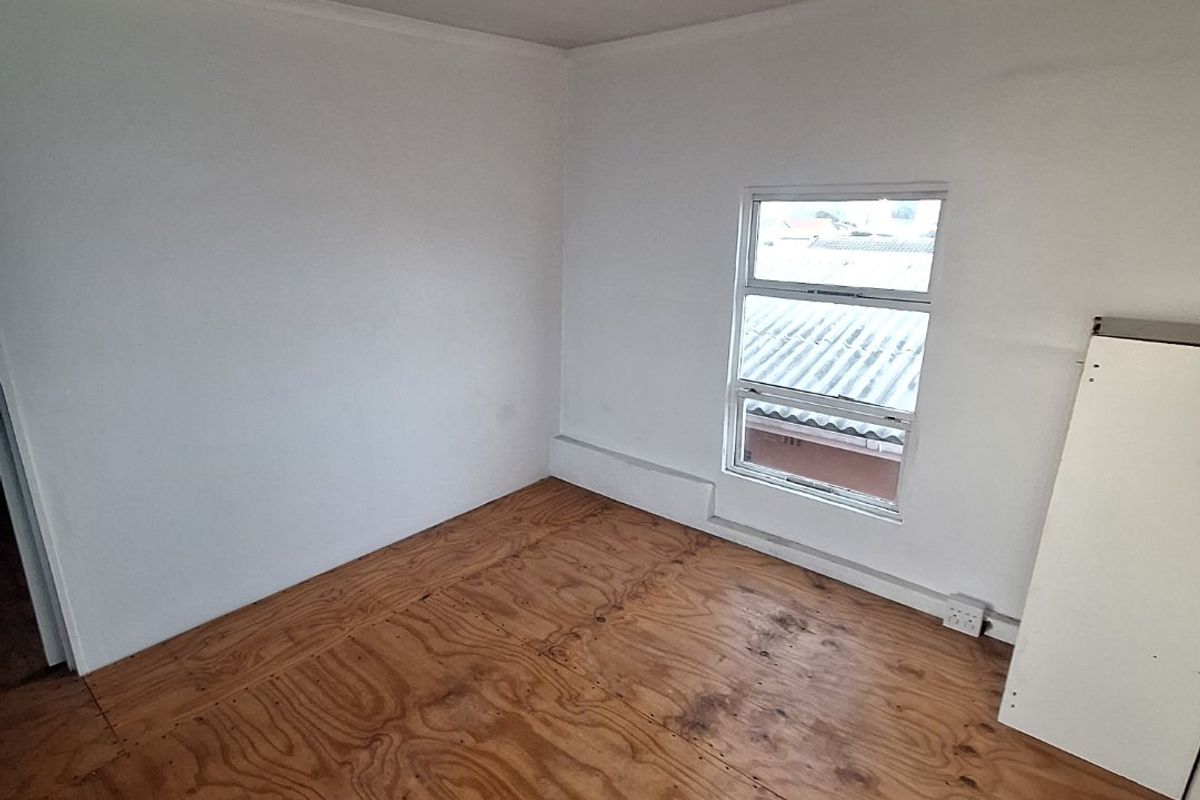 An empty bedroom featuring unfinished plywood flooring and a single aluminium window, requiring final flooring installation and cosmetic finishing.