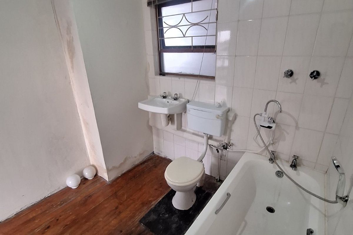 A dated bathroom featuring a bathtub, wall-mounted basin, and toilet, with visible wear on the walls and timber flooring requiring renovation.