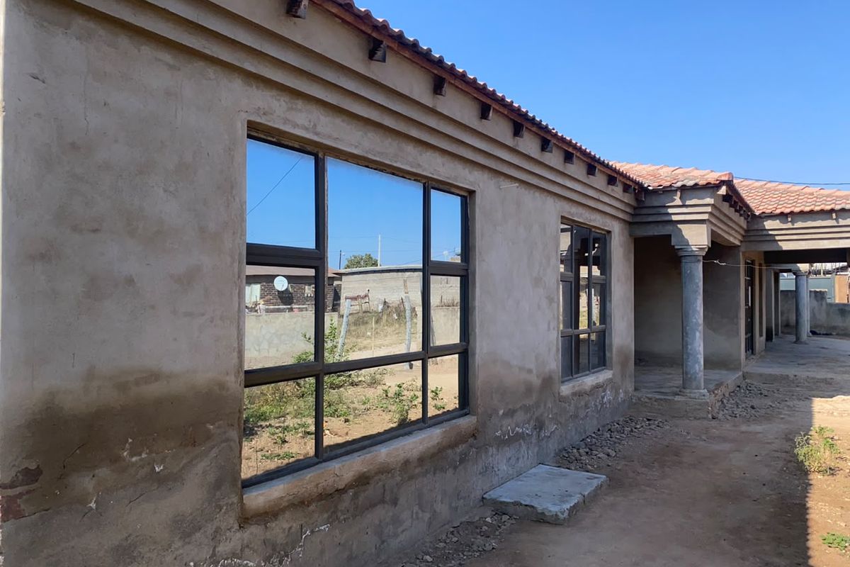 Rear exterior view of a house featuring unfinished plaster walls, aluminium window frames, and a tiled roof with concrete support columns.