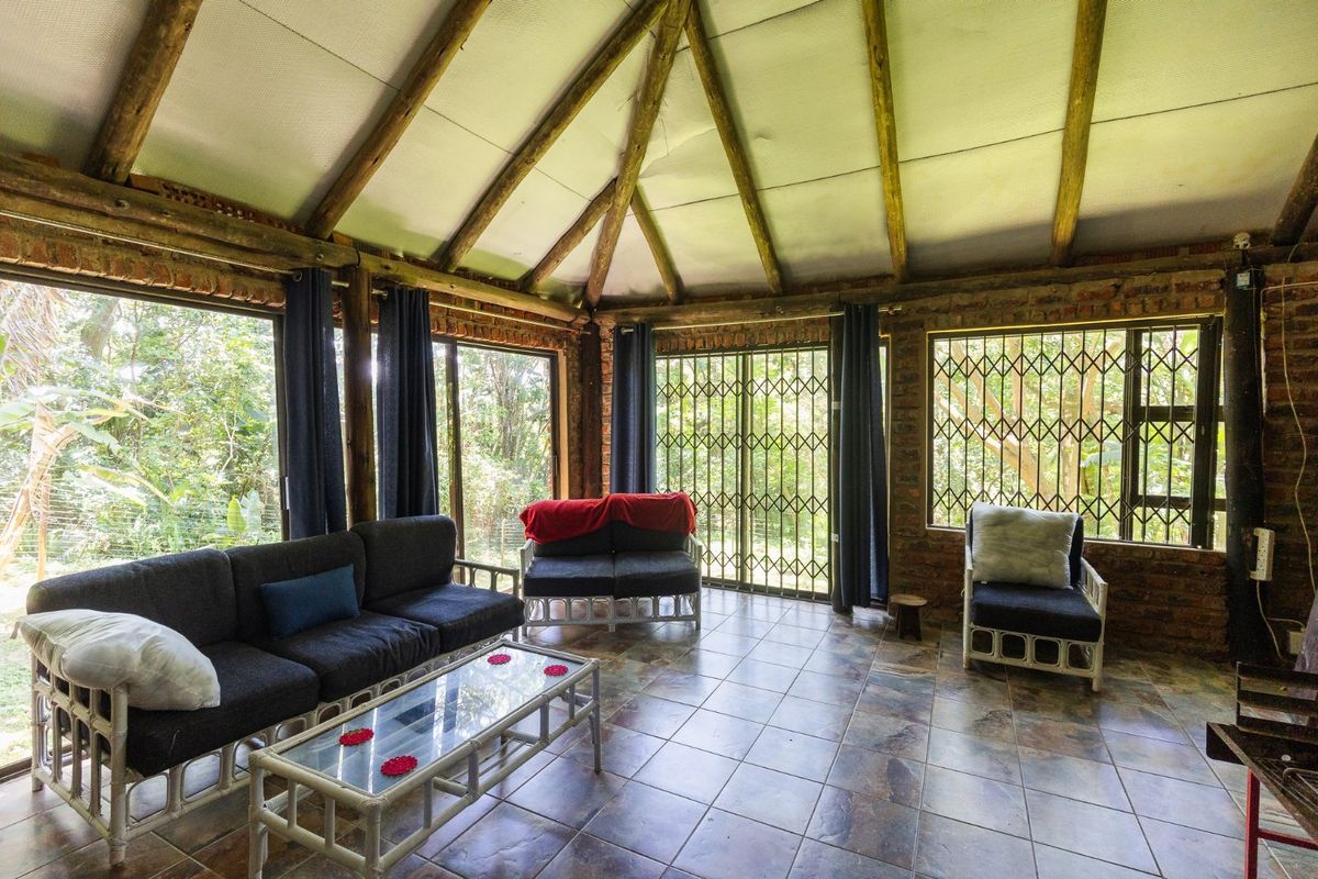 A rustic living room featuring exposed timber rafters, brick walls, and large sliding glass doors with security gates, offering views of the surrounding garden.