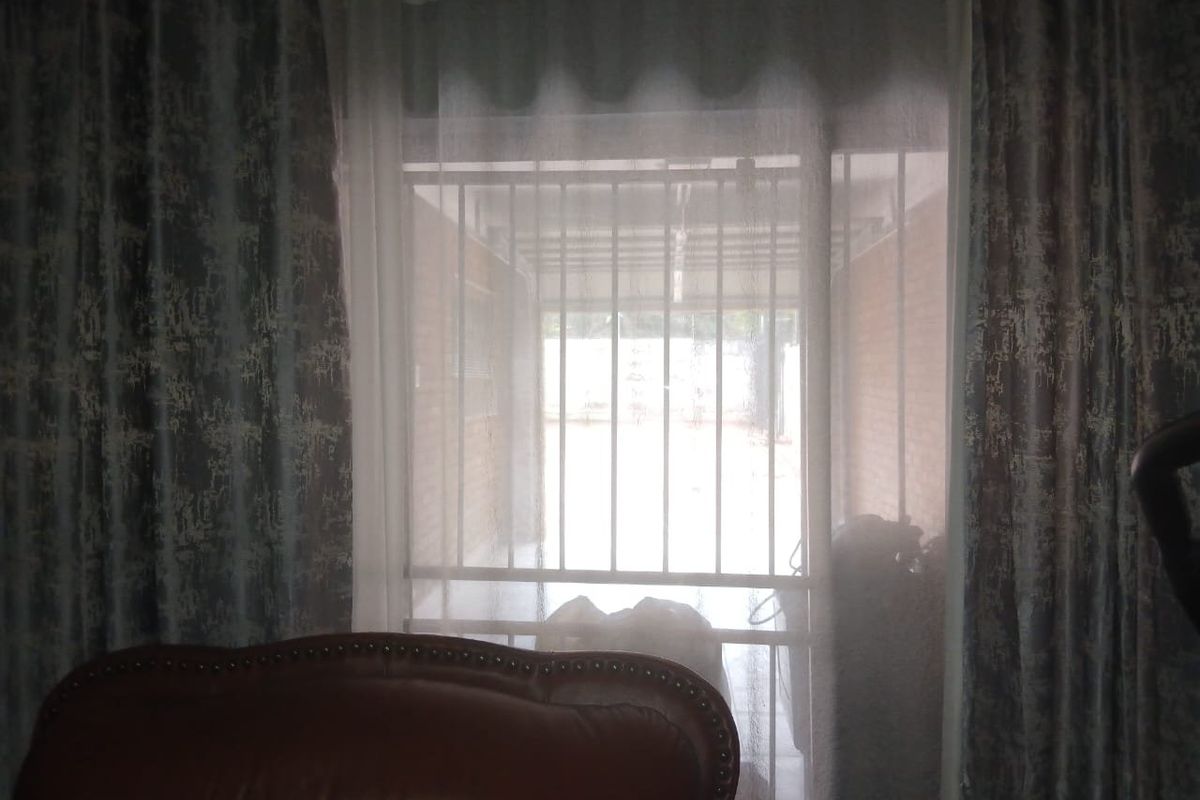 Interior view looking through a window with curtains towards an exterior security gate and covered outdoor area.