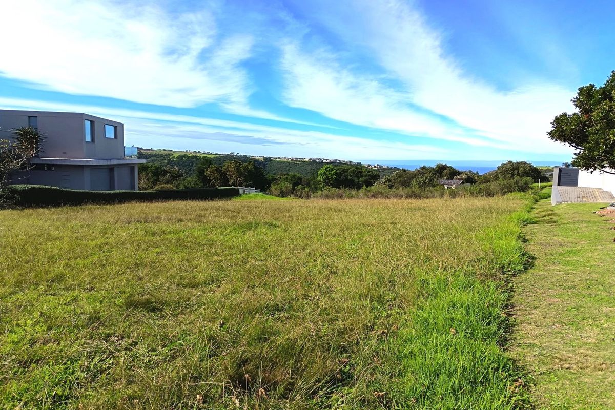 A vacant residential land plot featuring open grassy terrain with distant coastal views and adjacent modern residential architecture.