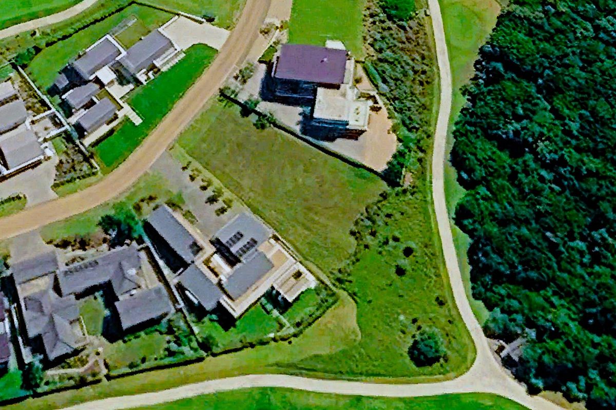Aerial view of a vacant residential plot situated within a developed estate, bordered by paved roads and natural forest vegetation.