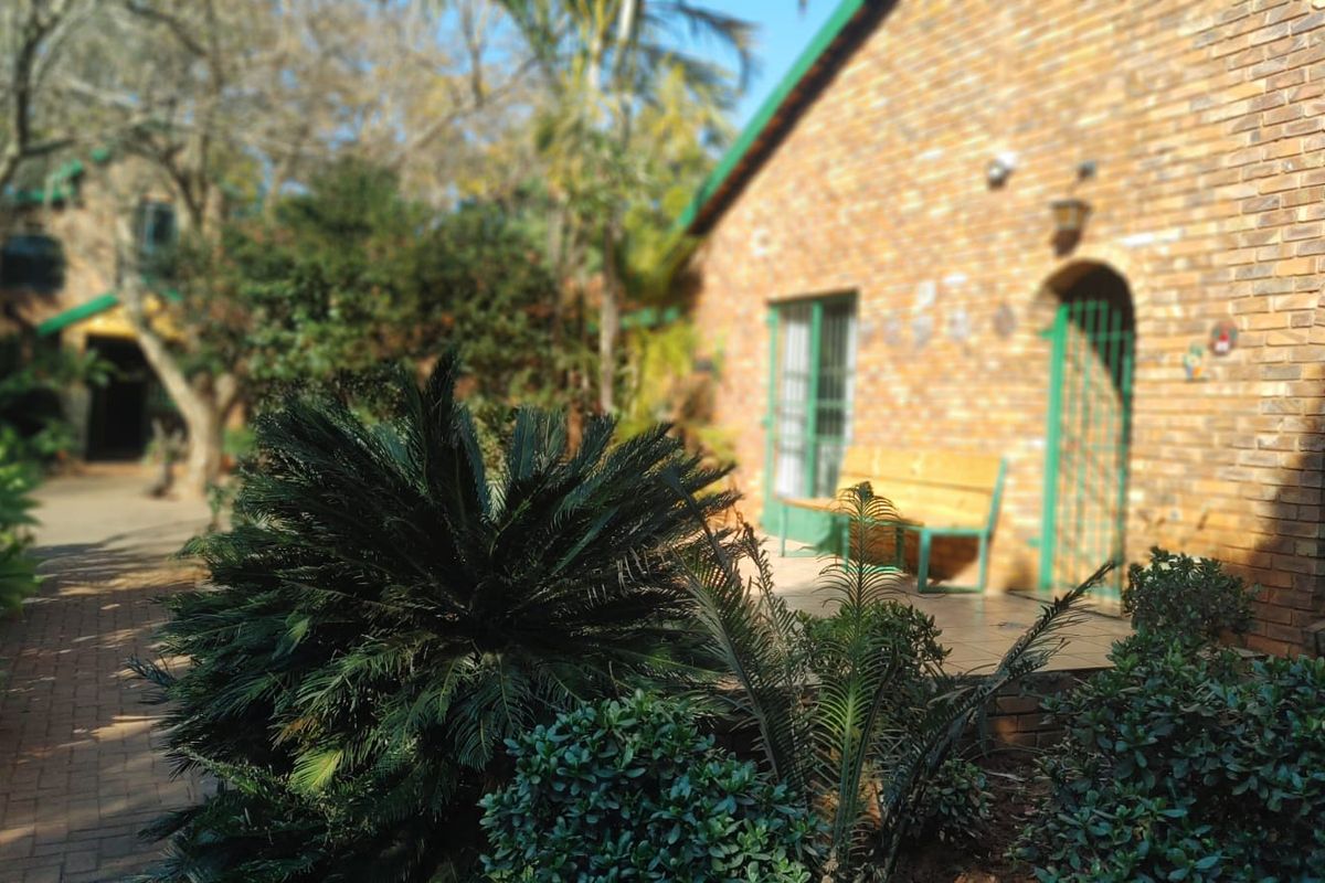 A rear view of the property featuring a face brick facade, a paved walkway, and a tiled patio area surrounded by established garden landscaping.