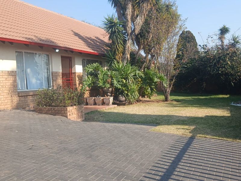 Front view of a single-story brick house featuring a tiled roof, paved driveway, and established garden area.