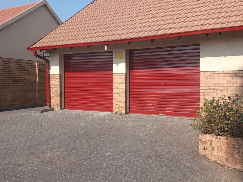 A double garage featuring red roller doors, a tiled roof, and a paved driveway with an integrated brick planter box.