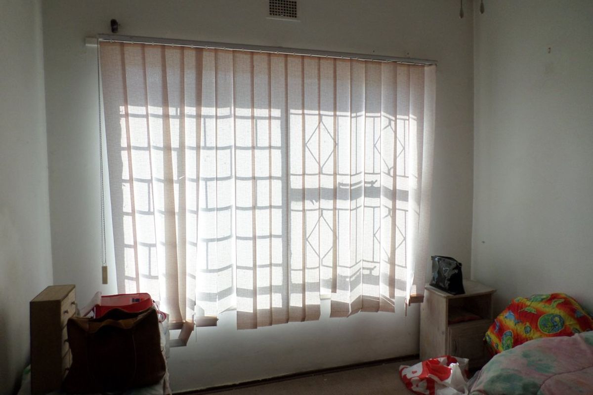 A bedroom featuring a window with security bars and vertical blinds, requiring cosmetic updates to the walls and flooring.