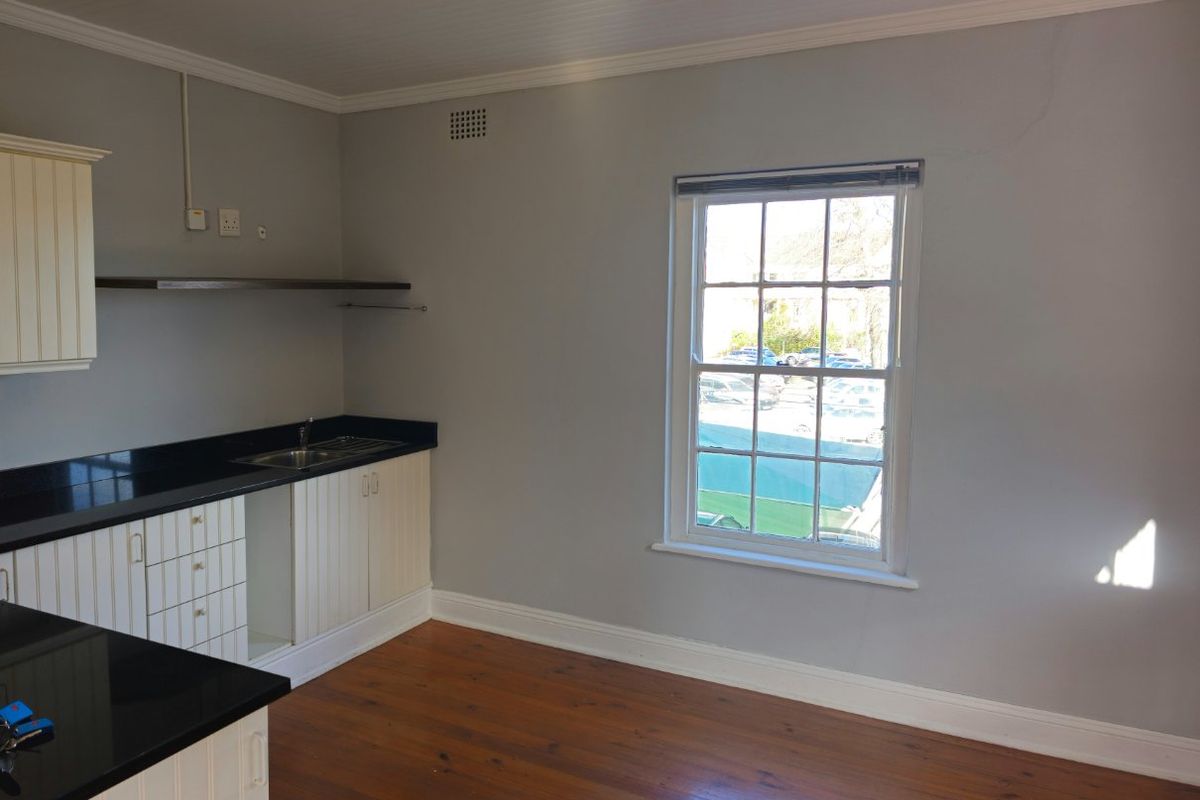A functional kitchen area featuring timber flooring, built-in cabinetry with black stone countertops, and a large sash window providing natural light.