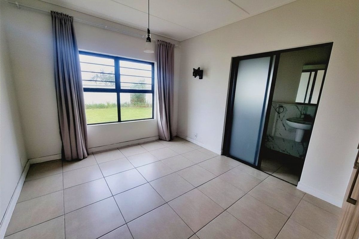A bright, empty bedroom featuring tiled flooring, a large window with security bars, and an adjoining en-suite bathroom with a sliding door.